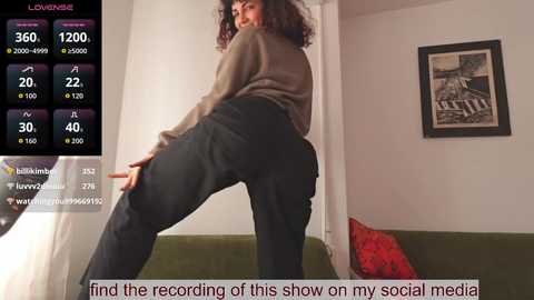 Video of a woman in a brown sweater, black pants, bending over, in a modern living room with a green couch, red pillow, and a framed photo on the wall.