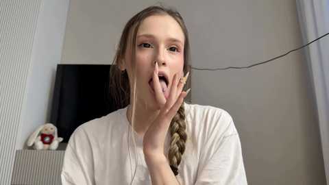 A young woman with light skin and long, blonde braided hair, wearing a white t-shirt, sticks out her tongue and covers her mouth with her hand. The background features a beige wall and a black TV with a teddy bear on a shelf.