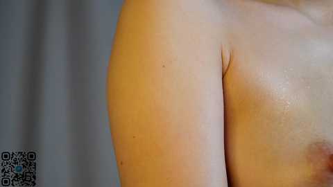Video of a close-up of a person's right shoulder and upper arm, showing a small mole and smooth, light skin. The background is blurred with a QR code in the bottom left corner.