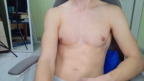 Video of a topless, fair-skinned, slender male with small breasts, seated in a grey chair, revealing a blue cloth on his lap. Background includes a mirror, green walls, and scattered objects.