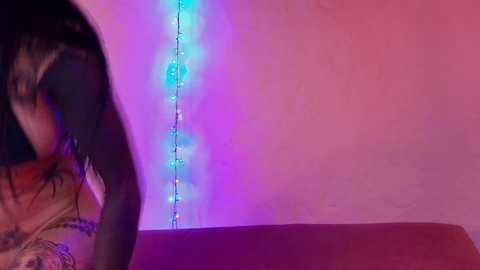 Media: Video of a woman with dark hair and tattoos, partially obscured by a pink wall with a blue LED string light.