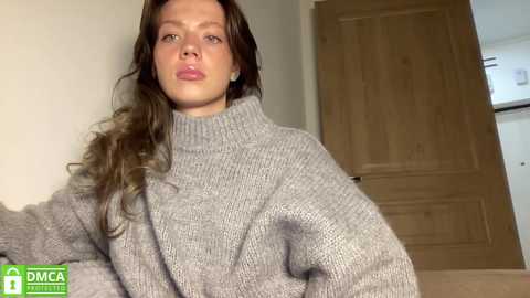 A video of a fair-skinned woman with long, wavy brown hair, wearing a gray knitted turtleneck sweater, standing indoors against a beige wall and wooden door.