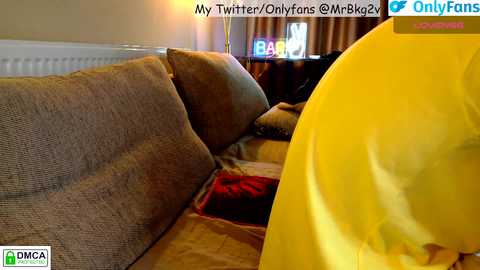 A video of a plush, brown sectional couch with a bright yellow shirt draped over it, set in a cozy, warmly lit living room with beige walls and a white radiator.