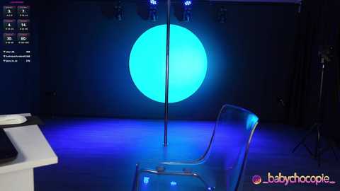 Media: Video of a dimly lit, modern stage with a large, glowing blue circle backdrop, a clear chair, and a spotlight.