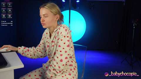 Media: Video of a young blonde woman in a red heart-patterned pajama set, seated at a white table, looking serene, with a blue light background and studio equipment visible.