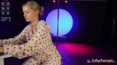 Media: Video of a blonde woman in a heart-patterned pajama set, sitting pensively at a table. Background features a large, circular, glowing blue light and a red floor.