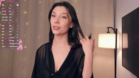 Video of a fair-skinned woman with long black hair, wearing a black sheer robe, standing in a softly lit room with a modern lamp and a flat-screen TV.