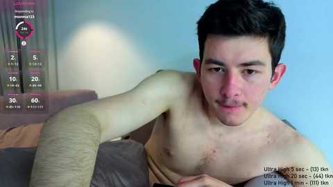 Media: Video of a young Asian man with short black hair, light skin, and a mustache, lying shirtless on a bed. He has a bandaged arm and a serious expression. The background shows a soft blue wall and a blurred window.