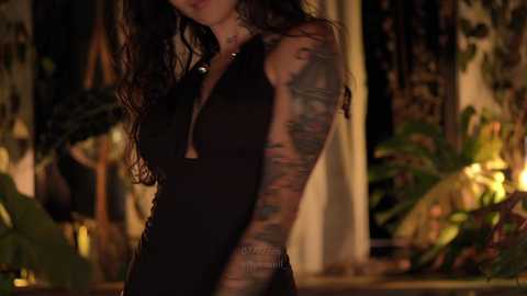 Media: Video of a woman with long, wavy dark hair, wearing a black sleeveless dress, showcasing her extensive colorful sleeve tattoos. Background features lush green plants and dim lighting.