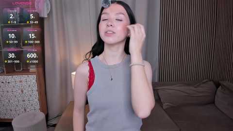 Video of a young woman with fair skin, long black hair, and a slender build, wearing a grey tank top and red bra, in a dimly lit bedroom with a wooden dresser and bed.