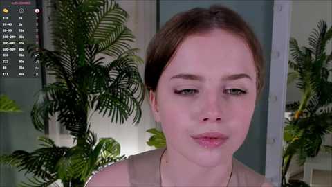 Video of a young Caucasian woman with fair skin and light brown hair, wearing a beige top, in a room with green potted plants and a calendar interface.