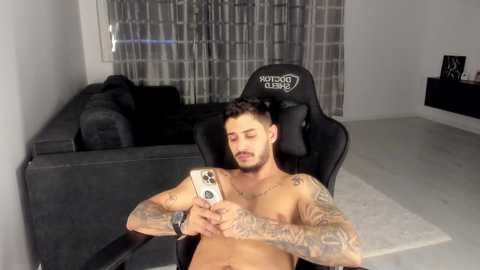 Video of a shirtless man with tattoos, seated in a black gaming chair, holding a phone, in a modern living room with a gray sectional and a white curtain backdrop.