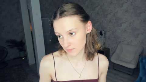 A video of a young, fair-skinned woman with shoulder-length brown hair, wearing a maroon spaghetti-strap top and a gold necklace, standing in a dimly-lit, modern room with a concrete wall and white chair.