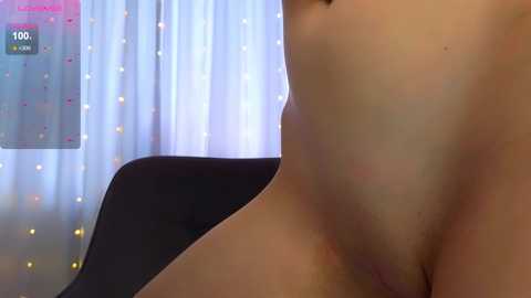 Video of a nude, light-skinned person with smooth skin, viewed from the side, showing the lower abdomen and partially obscured genital area. Background features a white curtain with small, colorful dots and a black chair.