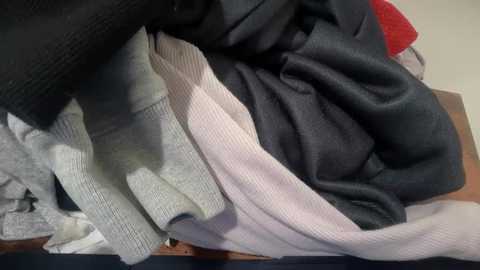 Video of a pile of gray, black, and white knit sweaters, one with a red collar, on a dark blue surface. The textures vary from soft to slightly rough.