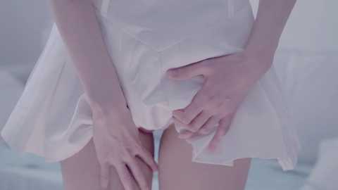 Video of a slender, light-skinned woman wearing a white, flowy dress, her hands gently lifting the skirt to reveal her bare thighs. Soft, pastel-toned background.