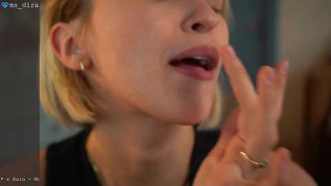 Video of a fair-skinned woman with blonde hair, wearing a black top, making a peace sign with her fingers. She has a small gold ring on her finger. Background is blurred.