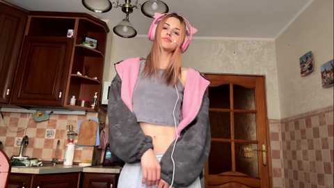 A video of a young woman with long, straight brown hair wearing pink cat ear headphones and a gray crop top, standing in a cozy kitchen with wooden cabinets, checkered wallpaper, and a wooden door.