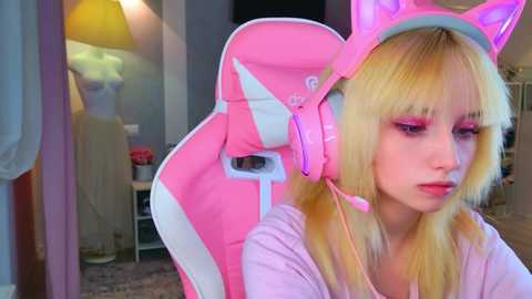 Video of a young woman with fair skin, wearing pink cat ears and headset, in a cozy room with a sewing machine and clothes in the background.