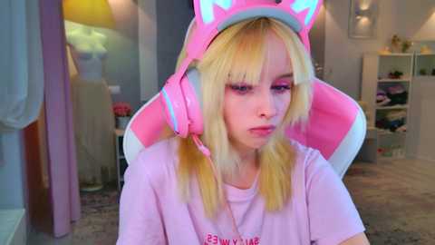Video of a young blonde woman in a pink gaming headset, wearing a light pink T-shirt, sitting in a cozy bedroom with a bed and shelves in the background.