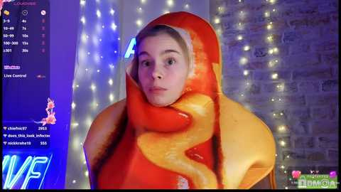 Media: Video of a young woman with fair skin and blonde hair, wearing a red, snake-like Halloween costume. The background features a dimly lit, brick-wall setting with string lights and a TV screen showing a live feed.