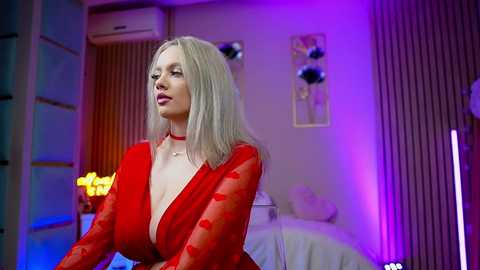 Video of a blonde woman with long hair, wearing a red, low-cut dress, in a modern bedroom with purple and blue lighting, visible air conditioning unit, and decorative wall art.