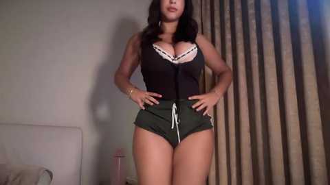 Video of a curvaceous woman with medium-dark skin tone, wearing a black lace-trimmed bra and olive green shorts, standing in a dimly lit bedroom with beige curtains and a bed visible.