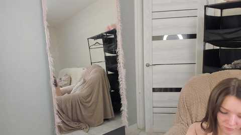 Video of a woman in a beige sweater, leaning against a mirror with a pink fringe, reflected in the mirror, showing a black clothing rack and a white door in the background.