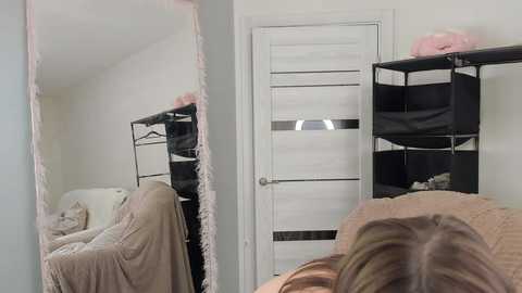 A video shows a person's back, blonde hair, and a reflection of a white door with a black handle in a small, minimalistic room.
