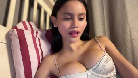 Video of an East Asian woman with long black hair, wearing a white lace bra, sitting on a bed with red and white striped pillows, looking seductive.