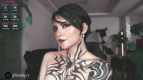 Media: Video of a pale-skinned, short-haired woman with dark makeup and extensive black tribal tattoos covering her chest, wearing a black top, in a dimly lit room.