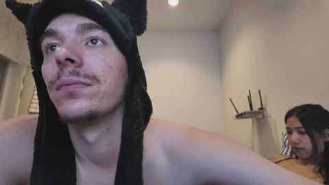 Video of a shirtless man with a black cat hat, looking serious, in a beige room with a woman in the background, holding a phone.