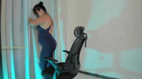 A video shows a woman with long black hair in a bun, wearing a navy blue backless bodysuit, standing in a dimly lit room with a black office chair, and a projector screen displaying text.