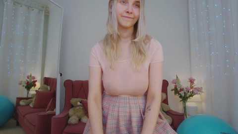 Media: Video of a young Caucasian woman with long blonde hair, wearing a pink t-shirt and plaid skirt, sitting on a red couch in a cozy room with pastel-colored decor and floral accents.