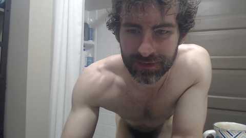 Video of a shirtless, muscular man with curly brown hair and a beard, looking directly at the camera, in a bathroom with beige walls and a white towel.