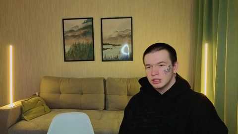 Video of a man in a black coat, with tattoos on his face, sitting on a beige sofa with a green cushion. Two framed landscape paintings hang on a beige wall, and green curtains are visible.