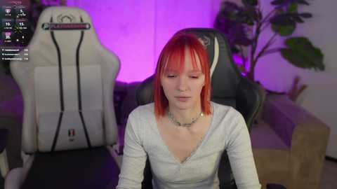 Video of a young woman with short red hair, wearing a white V-neck top, sitting in a gaming chair under purple lighting in a modern living room.