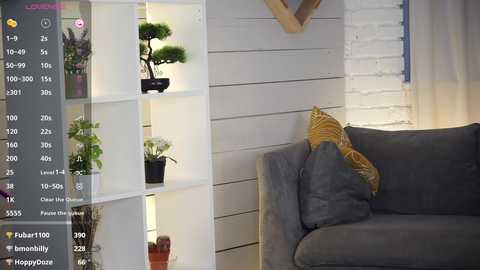 A video of a modern living room with a grey sofa, white brick wall, potted plants, and a light-filled shelving unit.