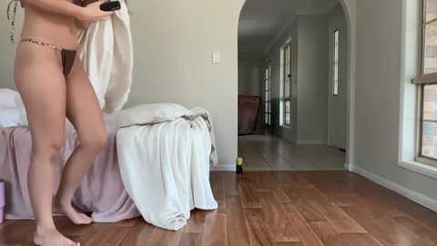 Video of a woman in a brown bikini, standing on a wooden floor in a bright, sparsely furnished room with white walls and an archway leading to another space.