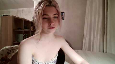 Video of a young, fair-skinned woman with blonde hair, wearing a floral-patterned strapless top, sitting on a bed in a softly lit, cozy bedroom.