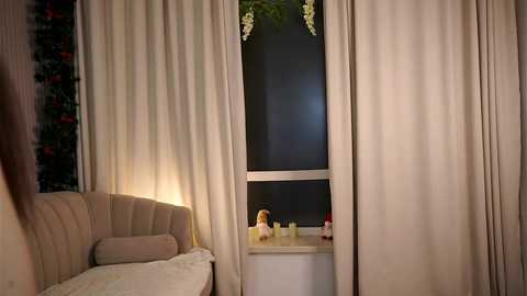 Media: Video of a cozy bedroom with beige curtains, a plush light pink sofa, a window with a small white shelf holding decorative items, and a soft yellow glow from a lamp.