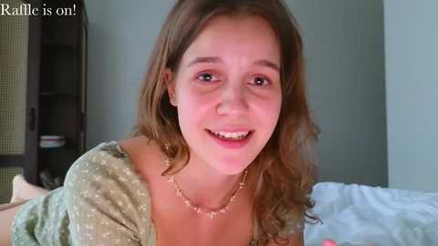 Video of a young Caucasian woman with wavy brown hair, smiling, wearing a green polka-dot blouse, in a bedroom with white sheets and a dark bookshelf in the background.