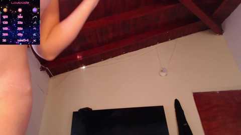 Video of a nude woman with fair skin and small breasts, standing in a room with a wooden ceiling, beige walls, and a flat-screen TV.