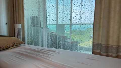 Video of a bedroom with a white bed, sheer curtains, a fan, and a view of a green landscape outside.