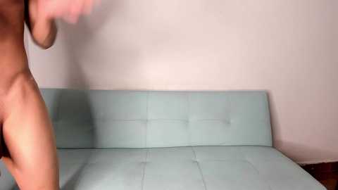 A video of a naked woman with fair skin and medium-sized breasts, bending forward in a minimalist room with a light blue couch against a white wall.