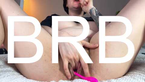 A video of a light-skinned woman with short hair, wearing a black shirt, using a pink vibrator on her shaved vulva. She has a tattoo on her arm. The background is blurred.