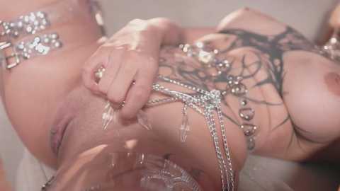 Video of a tattooed woman with fair skin, wearing intricate silver chains and a black lace bra, lying on her back.