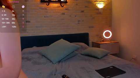 Video of a cozy bedroom with a blue upholstered bed, teal pillows, wooden nightstand, and a round mirror lamp, brick wall background, dim lighting.