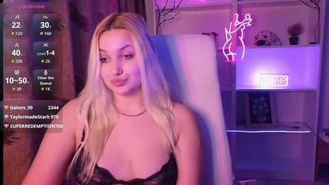 Video of a blonde woman with fair skin, wearing a black lace bra, sitting in a chair. Background features a purple-lit room with a neon sign, a clock, and shelves.