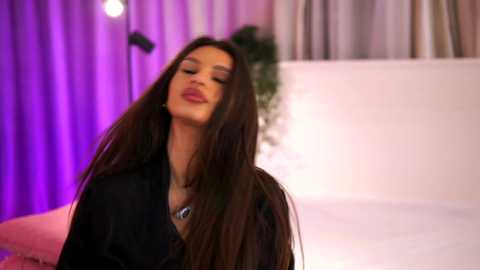 Video of a woman with long, dark hair, wearing a black shirt, standing in a brightly lit room with purple and white curtains and a potted plant.